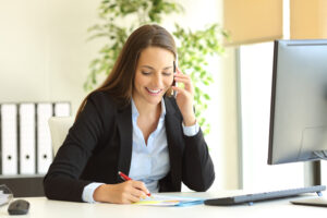 Happy businesswoman calling on mobile phone and taking notes on a desk at office